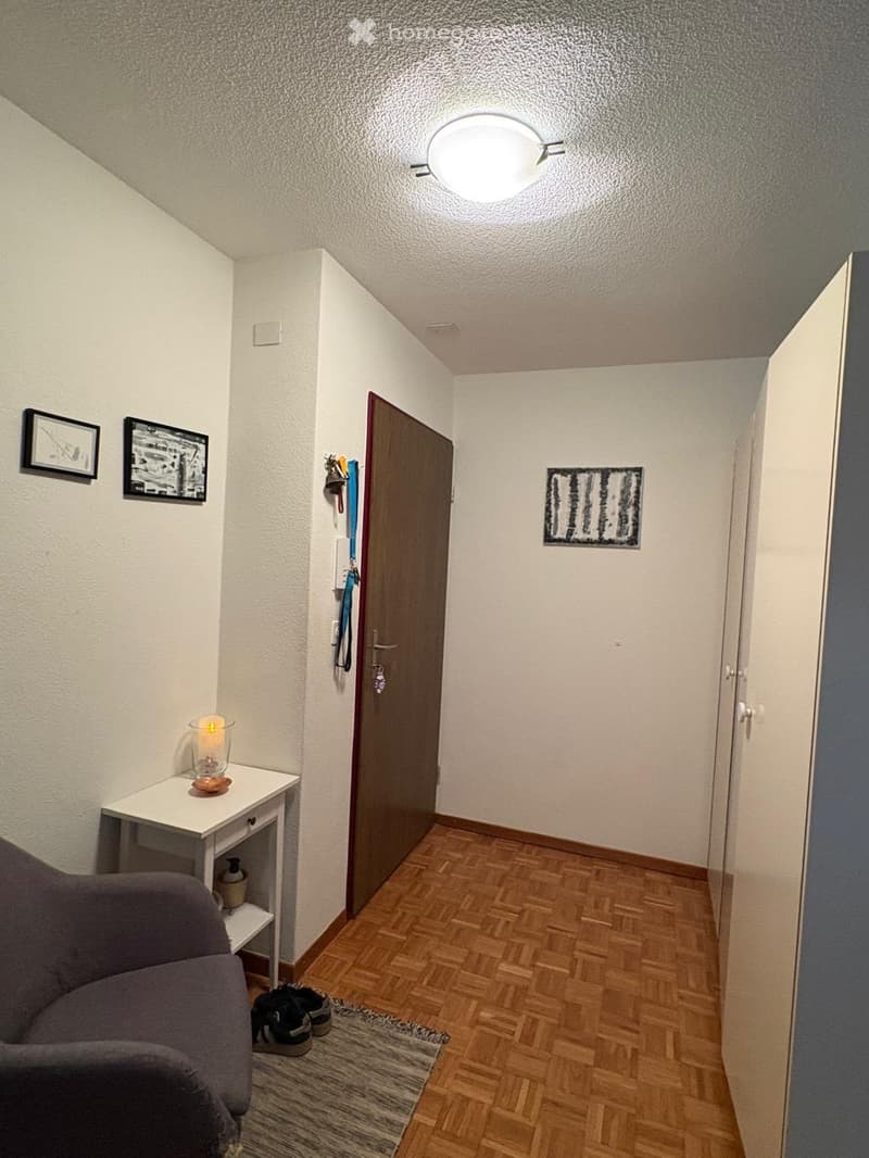 A small, well-lit hallway with wooden flooring, a grey sofa, a small white table with a candle, and a door leading to another room.
