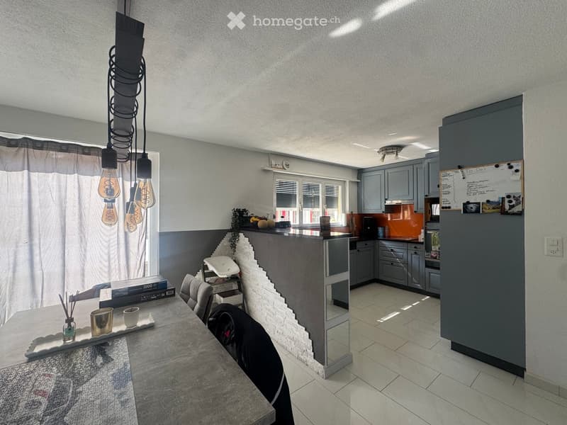 Modern kitchen and dining area with grey cabinets, a central island, and a dining table with chairs. Features include pendant lights, a window with blinds, and a whiteboard on the wall.