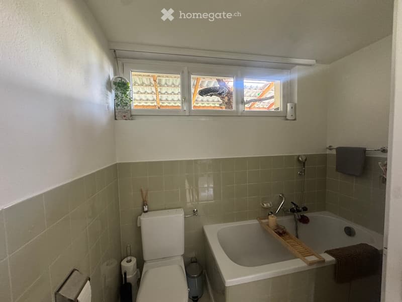 Bathroom with a toilet, bathtub, and a towel rack. The bathtub has a wooden tray with a candle and a small glass. The walls are tiled in a light green color, and there is a window above the bathtub.