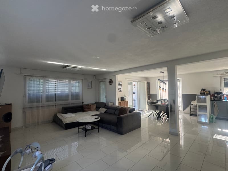 Modern living room with a large sectional sofa, coffee table, side table, dining area with table and chairs, and an open kitchen area with a counter and cabinets.