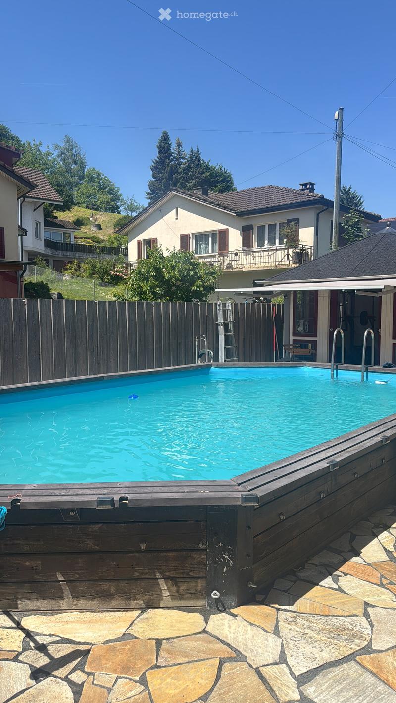 A swimming pool with a wooden deck, surrounded by a wooden fence, stone patio, and nearby residential houses with balconies and greenery.