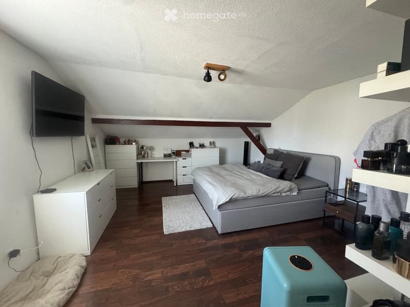 Cozy attic bedroom with wooden flooring, slanted ceiling, and modern furnishings including a bed, nightstand, desk, and shelves.