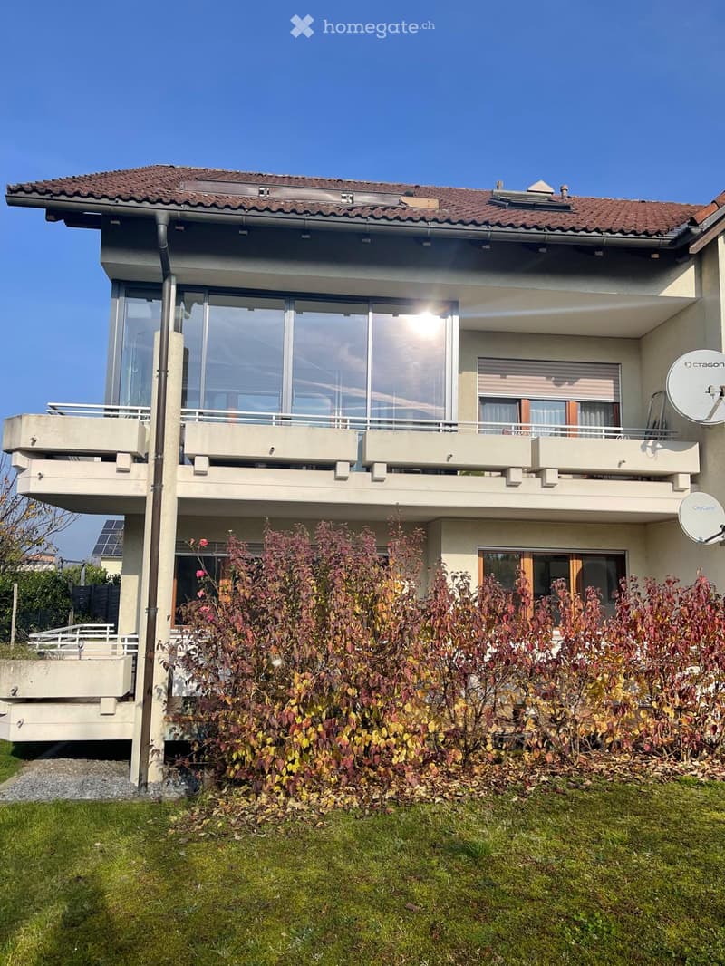 Two-story house with a tiled roof, large windows, balconies, and a well-maintained garden with shrubs and grass.