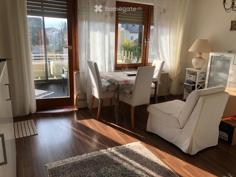 Bright living room with wooden flooring, large window with white curtains, sofa, armchair, dining table with chairs, side table with lamp, and candle holder on the windowsill.