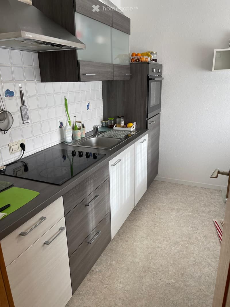 Modern kitchen with dark grey cabinets, white tiled backsplash, stainless steel sink, electric stovetop, and built-in oven. Countertop has a green cutting board and various kitchen utensils.