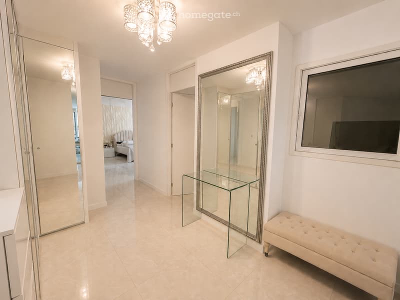 Elegant hallway with light-colored tiles, large mirrors, a glass coffee table, and a tufted bench. The hallway leads to a bedroom with a bed and nightstand visible in the background.