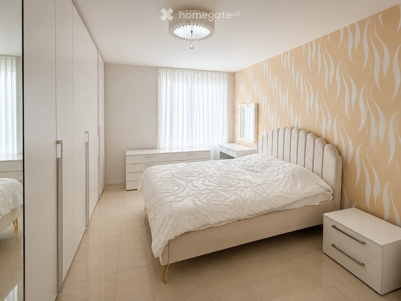 Modern bedroom with a large bed, white bedding, a nightstand, a mirror, and a wardrobe. The walls feature a decorative, flame-like pattern in warm tones.