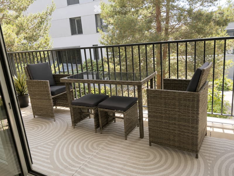 Balcony with wicker chairs, a glass-top table, two stools, and a potted plant, surrounded by a black metal railing and trees.