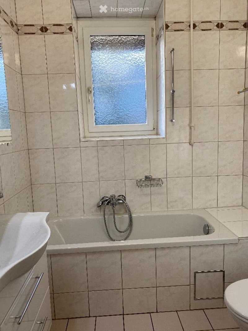 Bathroom with a bathtub, sink, toilet, and a window with frosted glass. The walls and floor are tiled in beige, and there is a towel rack and a shower hose visible.