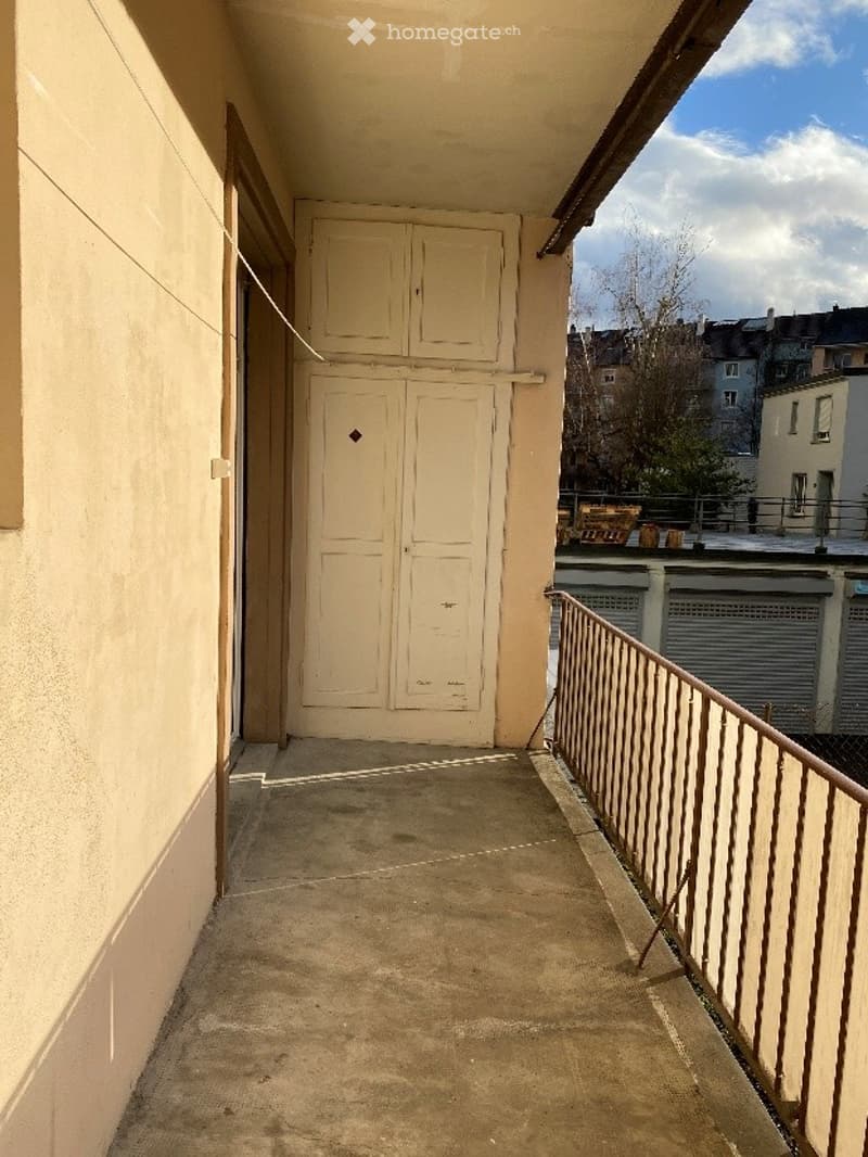 Balcony with a metal railing, a closed door leading inside, and a view of the city with buildings and trees.