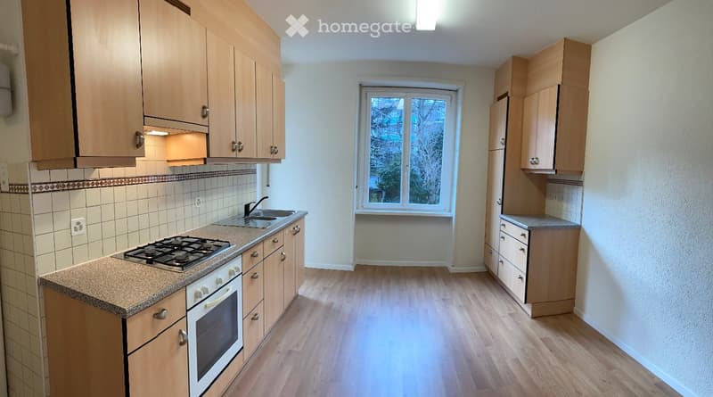 Bright kitchen with wooden cabinets, gas stove, oven, sink, and a window overlooking a tree-lined area.