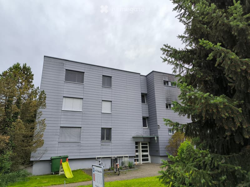 Modern two-story building with grey siding, multiple windows, and a small entrance area. There is a bicycle rack, a green trash bin, a yellow slide, and some trees surrounding the building.