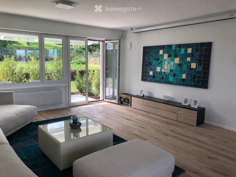 Modern living room with large windows offering a view of a garden, featuring a grey sectional sofa, glass coffee table, ottoman, wooden cabinet, and decorative artwork.