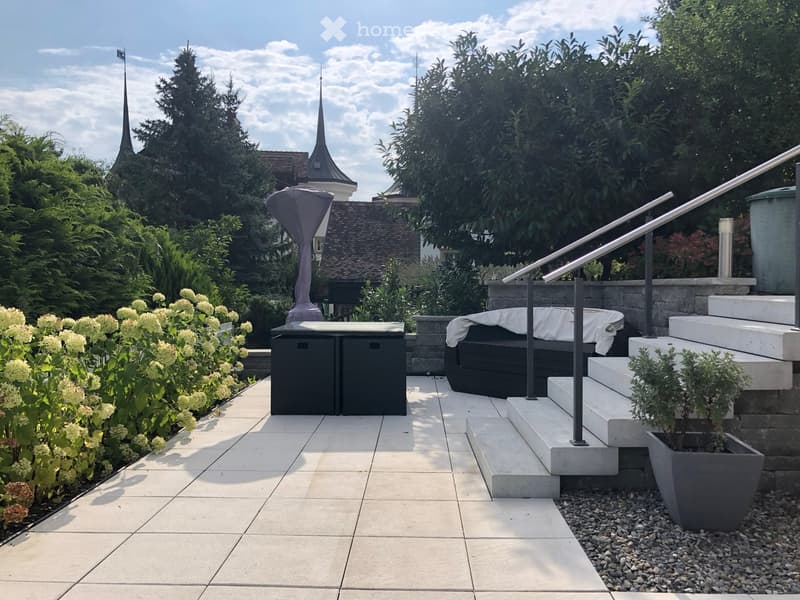 A well-maintained garden patio with a stone-tiled floor, a small staircase leading up, a black table and sofa set, a large umbrella, and lush greenery including flowering shrubs and trees.