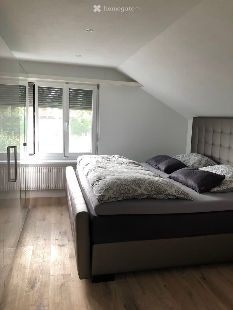 Modern bedroom with a large bed, patterned bedding, and dark pillows, featuring wooden flooring and large windows with blinds.