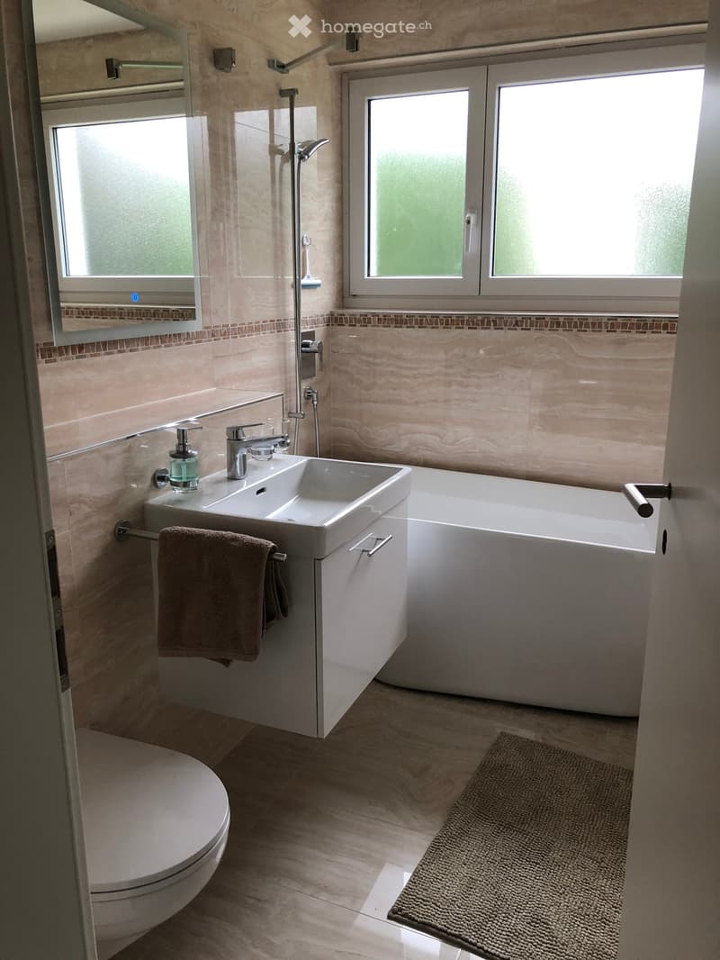 Modern bathroom with a white sink, bathtub, toilet, and a towel hanging on a rack. The walls are tiled in a light beige color with a decorative border, and the floor is tiled in a similar tone. A small bathroom rug is placed near the bathtub.