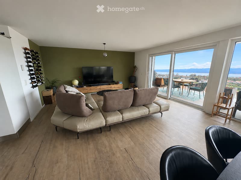 Modern living room with a sectional sofa, coffee table, television, and large windows offering a scenic view. Includes a wine rack, potted plant, and outdoor dining area visible through the glass doors.