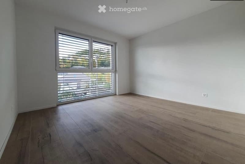 Empty room with wooden flooring, white walls, and large window with blinds.