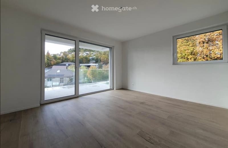 Empty room with wooden flooring, large sliding glass doors, and a window showing a view of trees and houses outside.