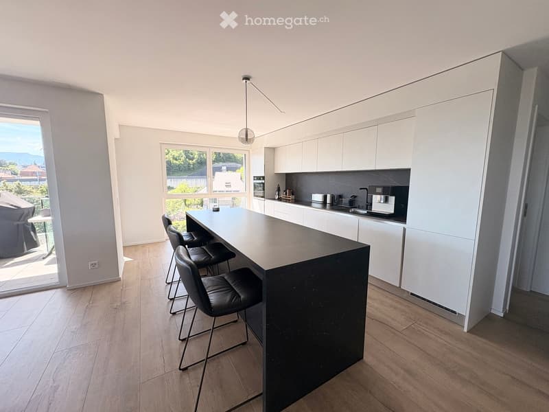Modern kitchen with white cabinets, a black kitchen island, bar stools, and various appliances including a sink, oven, microwave, toaster, and coffee maker. Large windows provide natural light and a view of the outdoors.