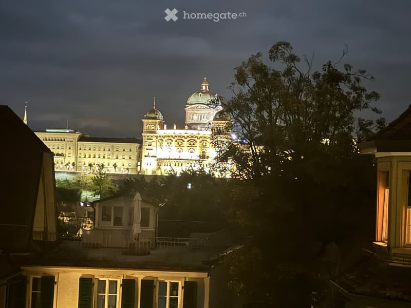 Nighttime view from a balcony or terrace showing a grand illuminated building with domes and spires in the background, surrounded by trees and smaller structures.