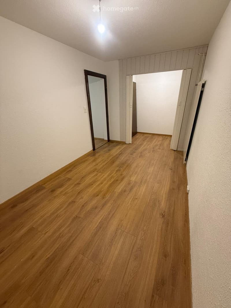 Empty hallway with wooden flooring, white walls, and a mirror on one side. The hallway leads to an open doorway.