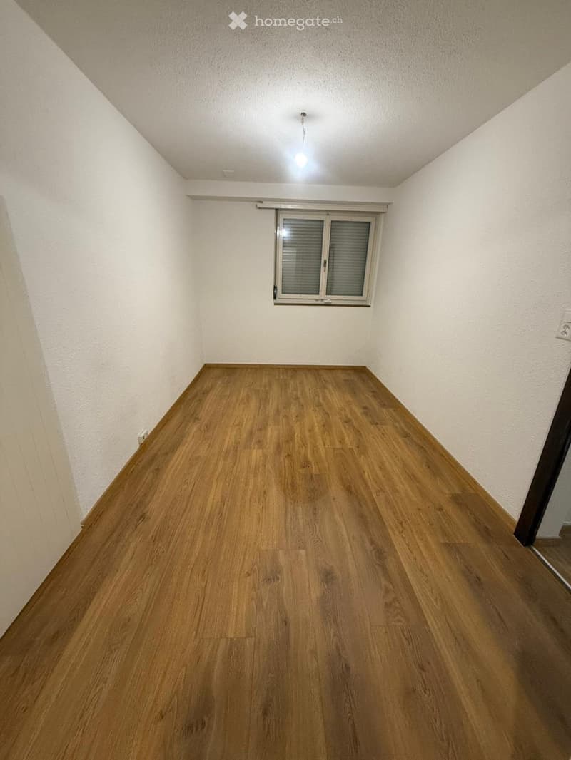 Empty room with wooden flooring, white walls, and a single window with blinds. The ceiling has a recessed light fixture.