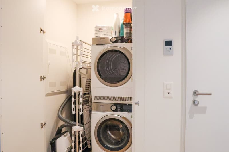 A compact laundry area with a stacked washing machine and dryer, a vacuum cleaner, and a storage rack with various items.