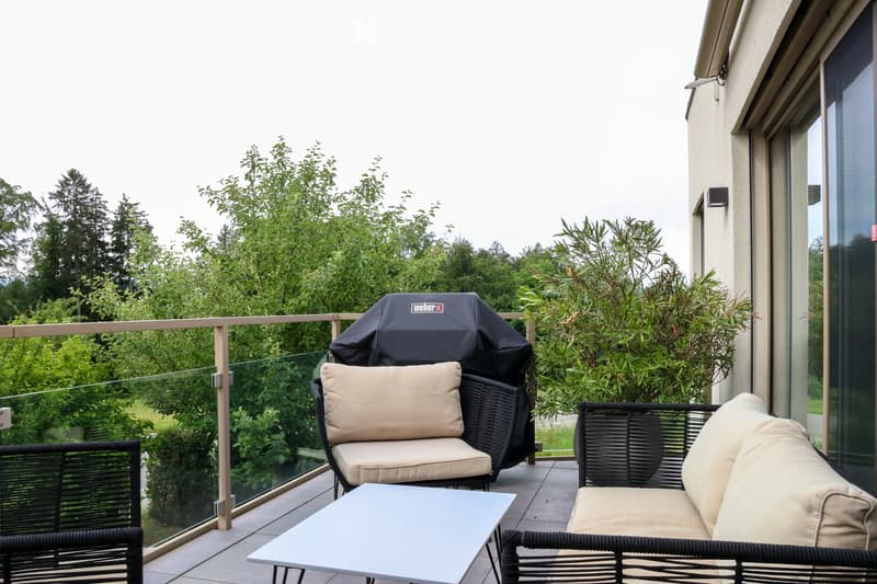 Balcony with a glass railing, featuring a small table, two wicker chairs with beige cushions, and a covered grill.