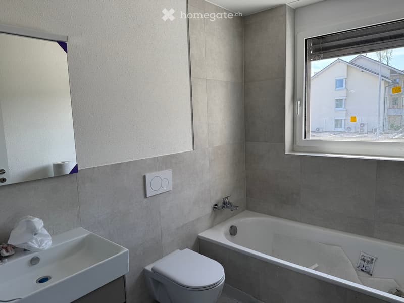 Modern bathroom with a white sink, toilet, bathtub, and mirror. The walls are tiled in a light grey color, and there is a window showing a view of neighboring buildings.