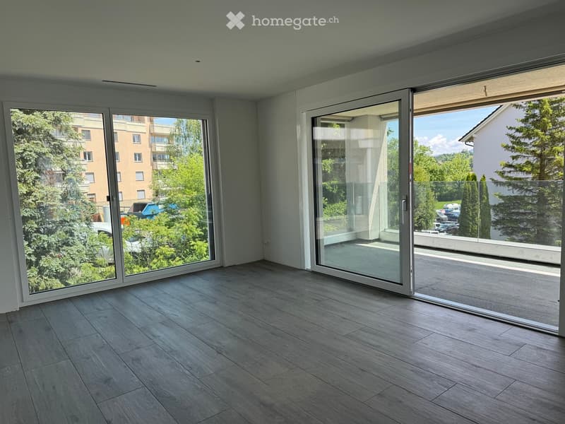 Empty room with light wooden flooring and large glass sliding doors leading to a balcony area with a view of greenery and buildings.