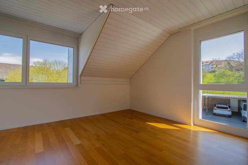 Bright, empty room with wooden flooring and slanted white ceiling. Large windows offer views of greenery and a nearby garage with parked cars.