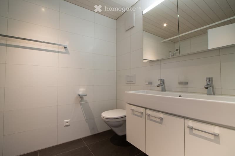 Modern bathroom with white tiles, a toilet, a double sink with chrome faucets, a large mirror, and white cabinets with silver handles.