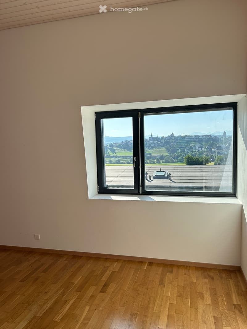 Empty room with wooden flooring, large window offering a scenic view of a cityscape and landscape.