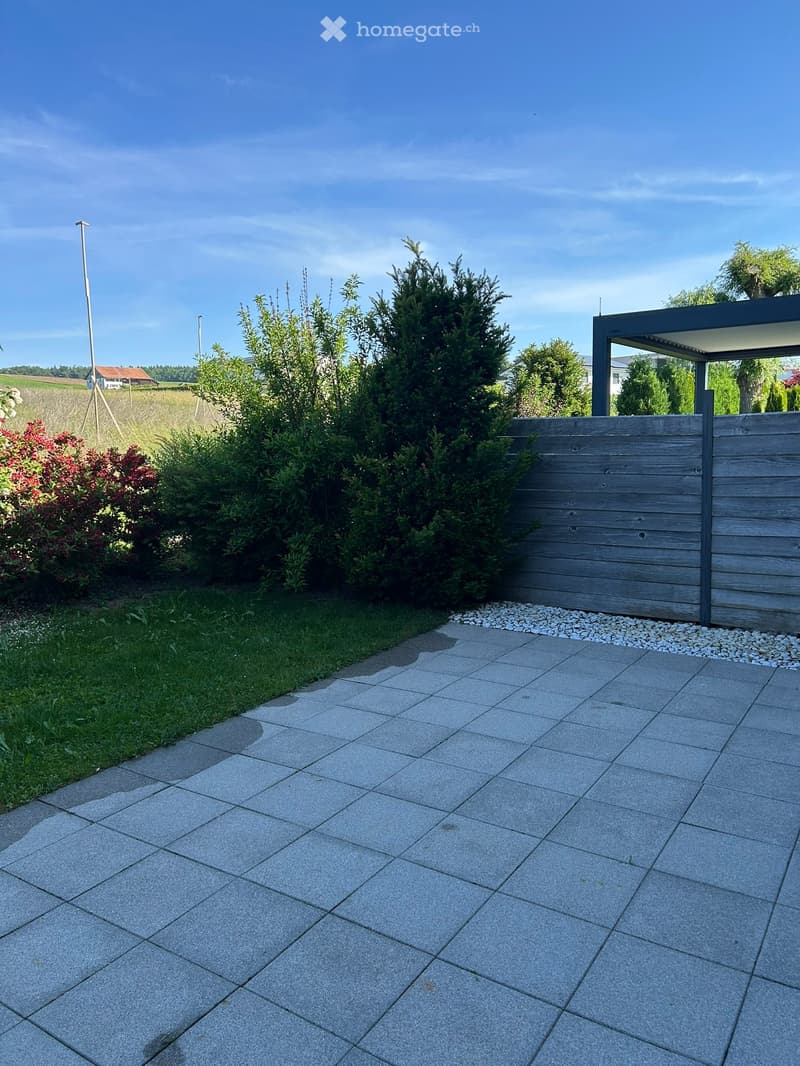Garden area with a paved patio, wooden fence, and various shrubs and trees.