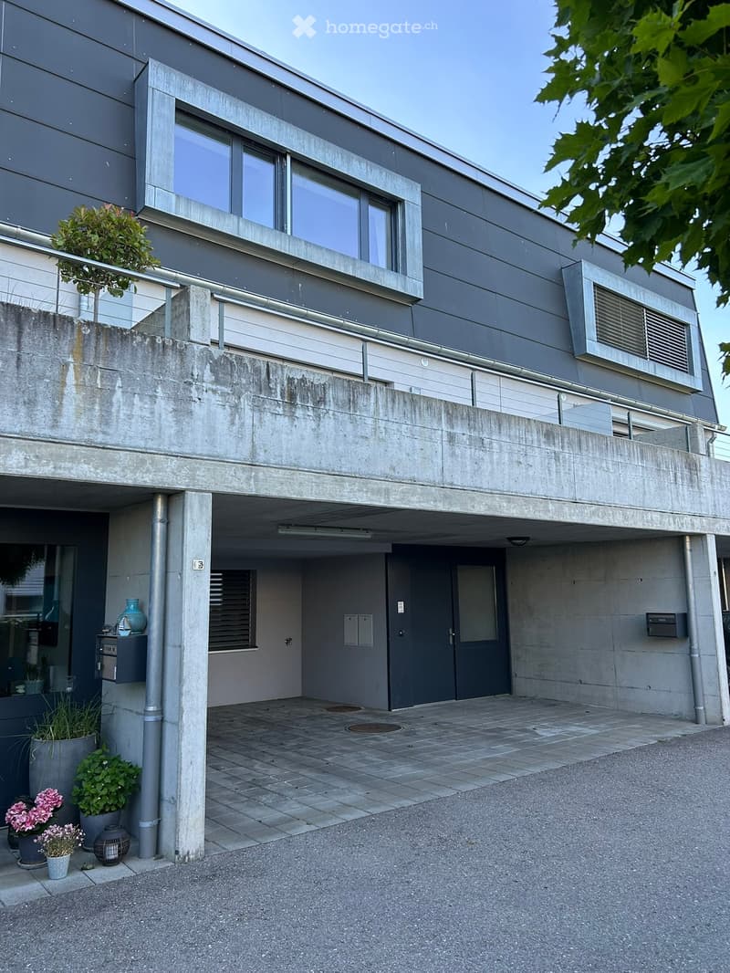 Modern two-story building with grey facade, raised ground floor, and balcony on the upper level. Features include a concrete entrance area with potted plants and a vase.