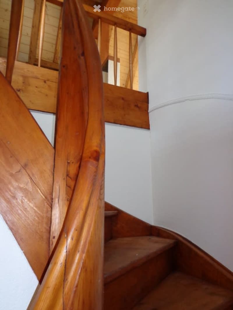 Spiral staircase with wooden handrails and steps, leading to an upper level with visible wooden railings and white walls.
