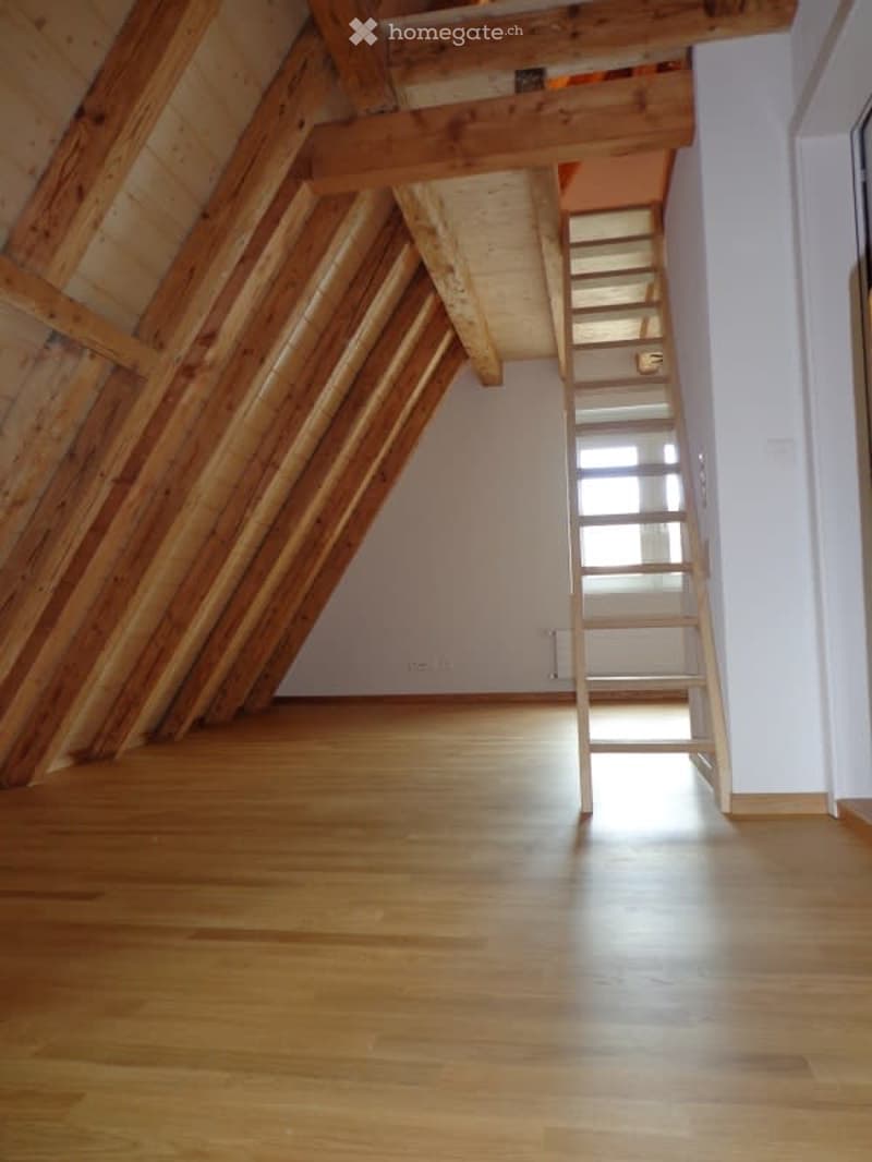 Empty attic space with wooden beams, wooden floor, and a narrow staircase leading up.