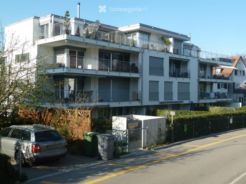 Modern multi-story apartment building with balconies, white exterior, and surrounding greenery.