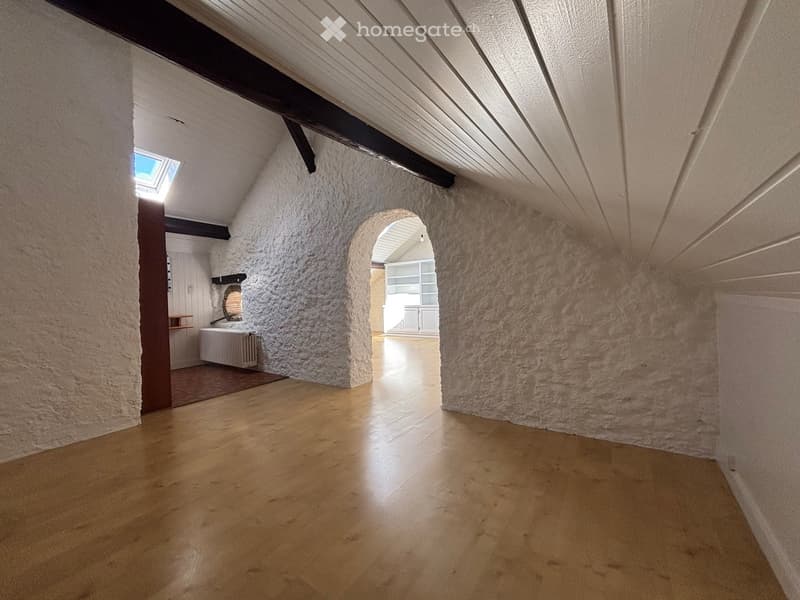 Empty room with wooden flooring, textured white walls, and exposed wooden beams on the ceiling. Features an arched doorway leading to another room and a skylight above.