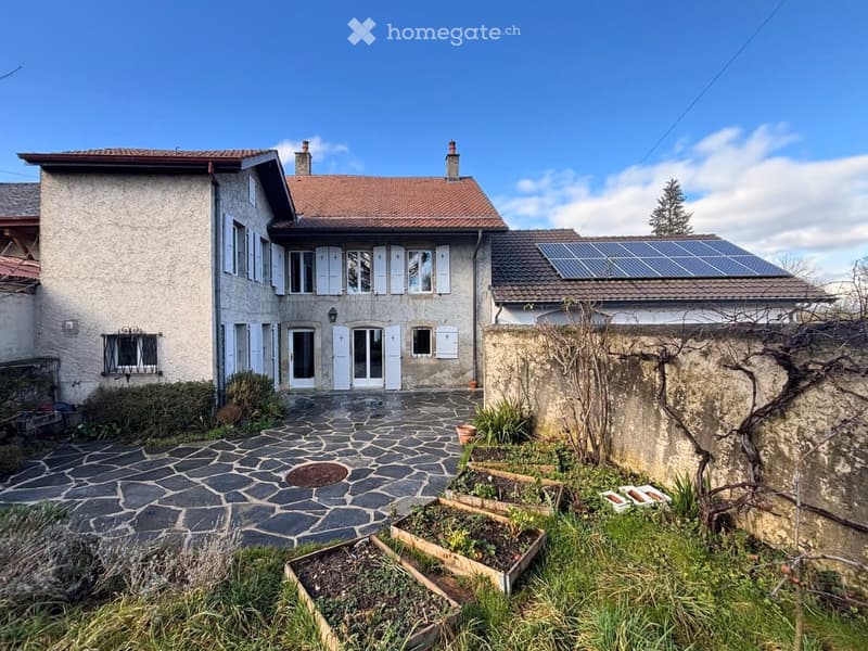 Two-story house with a red-tiled roof, white shutters, and solar panels on one section of the roof. The entrance features a stone-paved pathway leading to a glass door, surrounded by a small garden area with raised beds and a stone wall.