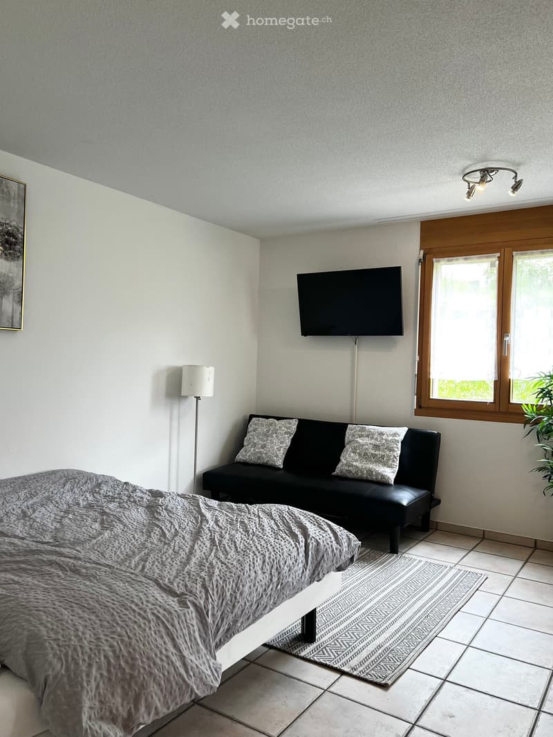 A modern living space with a bed, sofa, television, floor lamp, and a small table. The room features tiled flooring and large windows with wooden frames.