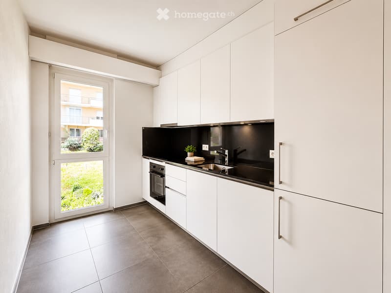 Modern kitchen with white cabinets, stainless steel sink, oven, and a small potted plant on the counter. The space features large windows offering a view of greenery outside.