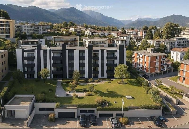 Aerial view of a modern residential complex with multiple apartment buildings, a central green courtyard, and a parking area with several cars.
