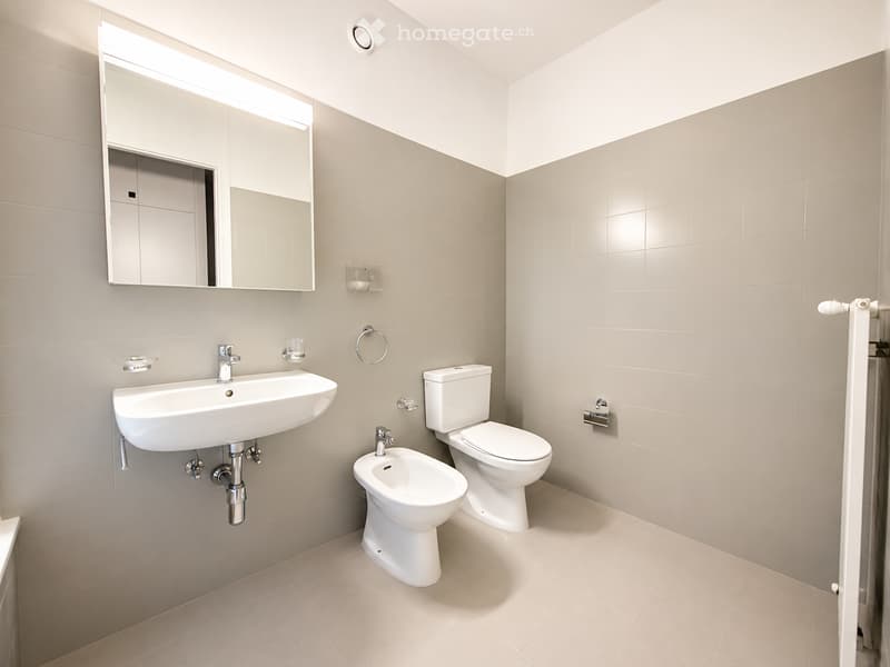 Modern bathroom with beige tiles, white fixtures including a sink, toilet, bidet, and a large mirror with built-in storage.