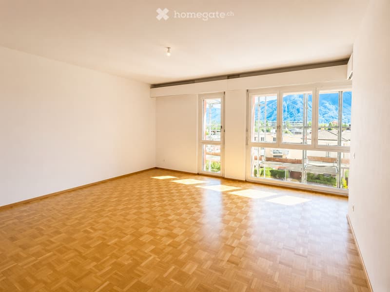 Empty room with wooden herringbone flooring, white walls, and large windows offering a scenic mountain view.