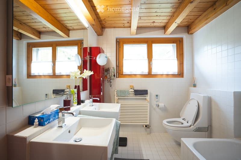 Bright bathroom with wooden ceiling beams, two sinks, a toilet, a bathtub, and a large mirror. The space features white tiles, a radiator, and decorative elements like a flower vase and towels.