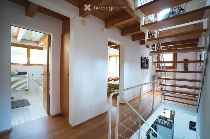 Bright hallway with wooden floors and exposed wooden beams. Features a staircase leading to an upper level, with visible rooms including a bathroom and bedroom.