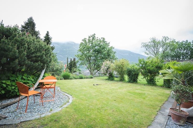 Well-maintained garden with a lawn, gravel pathway, outdoor seating area with a table and chairs, surrounded by trees and shrubs, with a scenic mountain view in the background.