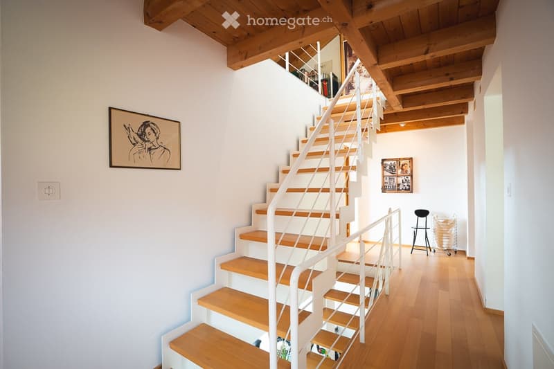 Modern hallway with wooden flooring, white walls, and a staircase leading to an upper level. Features include exposed wooden beams on the ceiling, a framed artwork on the wall, and a small black chair near the staircase.